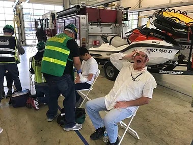 Emergency responders assist a man with a head injury, seated indoors near rescue equipment and a jet ski.