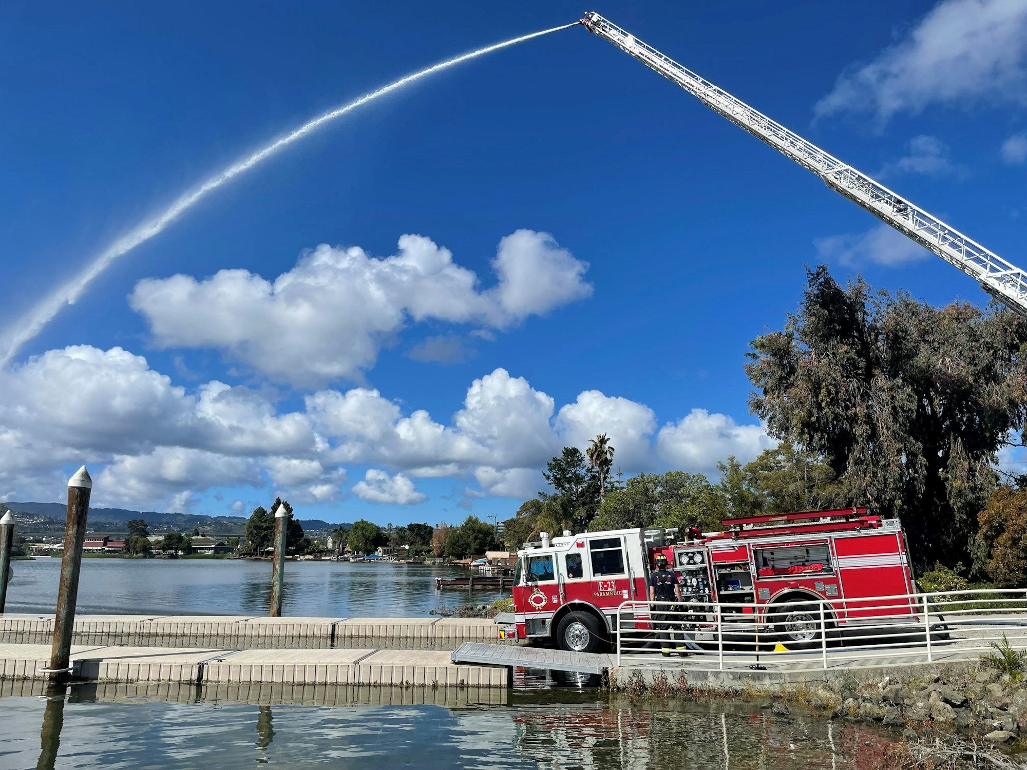 A fire truck by the water sprays a high arc of water into the sky on a sunny day with scattered clouds.