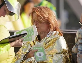 A person wrapped in a foil blanket holds a triage tag, while someone else writes on a clipboard.