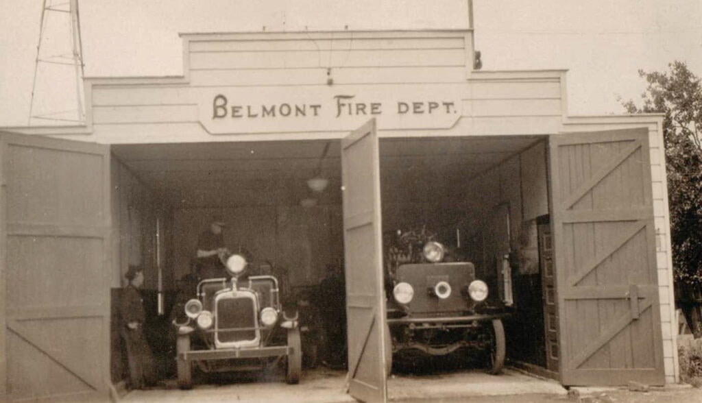 Vintage photo of two early fire trucks in a garage marked "Belmont Fire Dept." with open doors and people nearby.