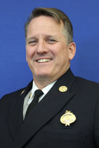A person in a uniform with badges on the jacket, smiling in front of a blue background.