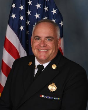 A person in a formal uniform with medals, smiling in front of a U.S. flag.