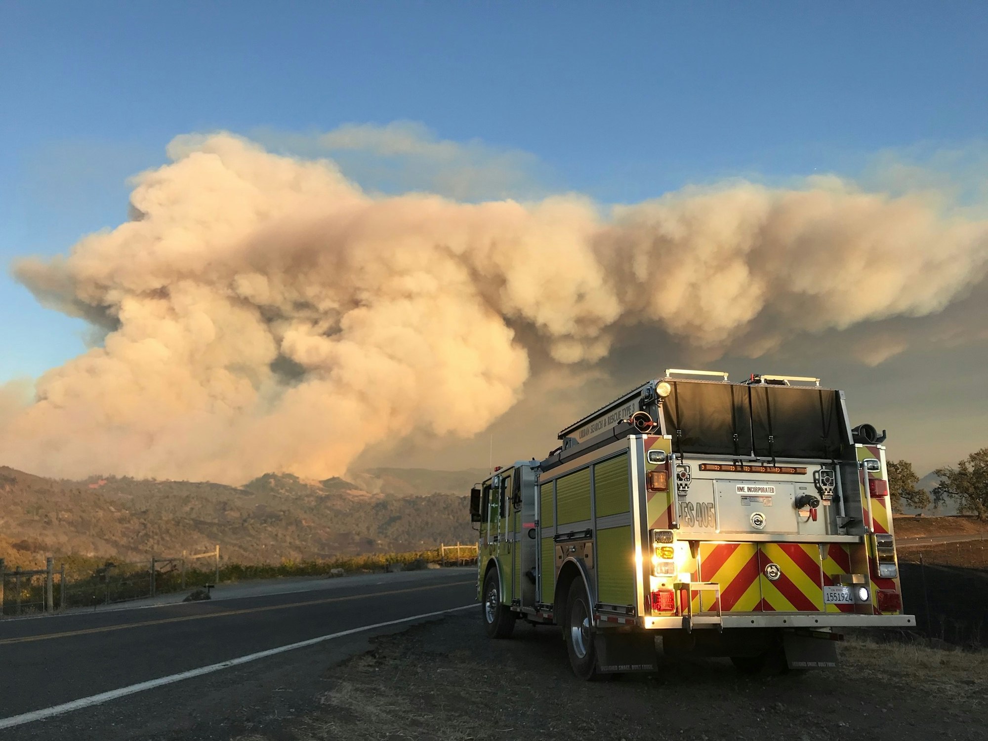 Fire truck on roadside, large smoke plume rises in the background, likely a wildfire in a scenic hilly area under clear skies.