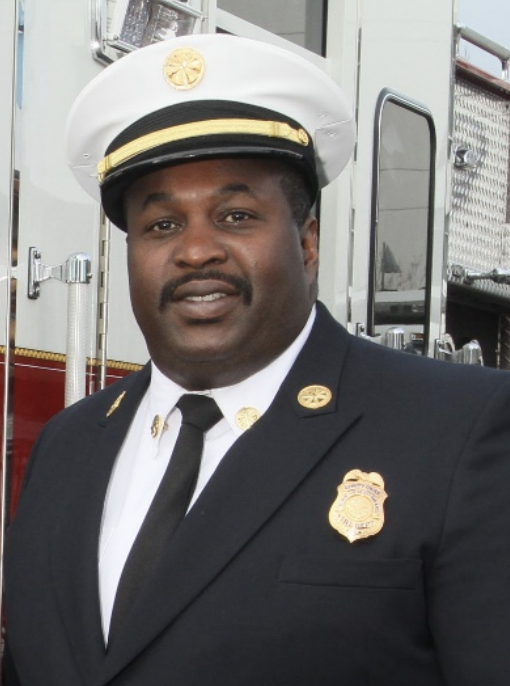 A person in a formal firefighter uniform, standing beside a fire truck.