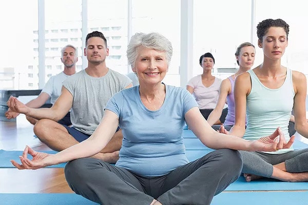 A group of people meditating on yoga mats, with a smiling elderly woman at the front.