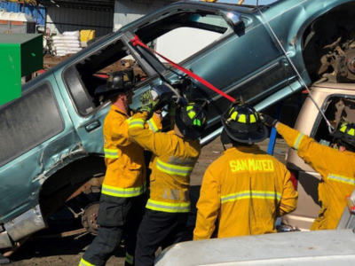 Firefighters in yellow jackets using tools to stabilize a tilted car at a rescue training scene.