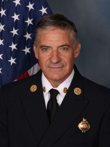 A person in a formal uniform with badges, standing in front of the American flag.