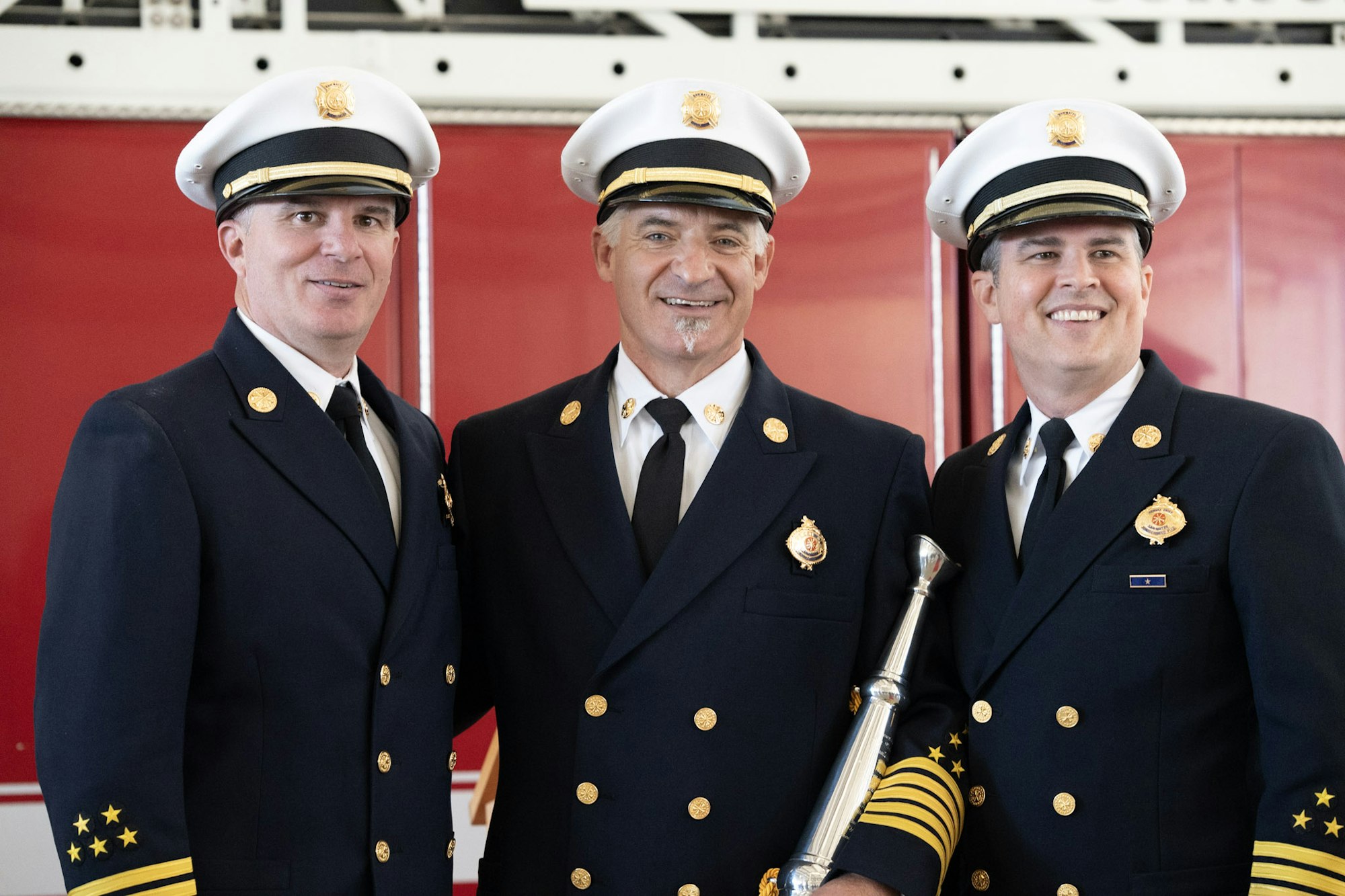Three men in uniform, wearing white peaked hats, pose in front of a red background.