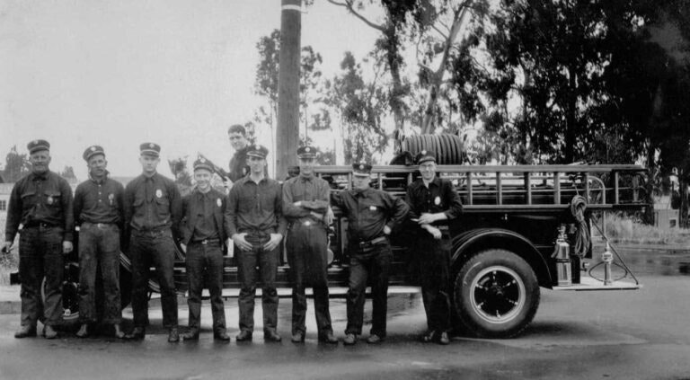 A group of firefighters in uniform standing in front of a vintage fire truck.