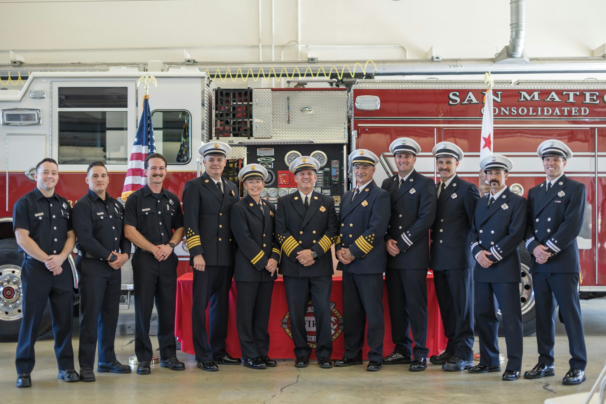 A group of firefighters in uniform posing in front of a fire truck with flags.