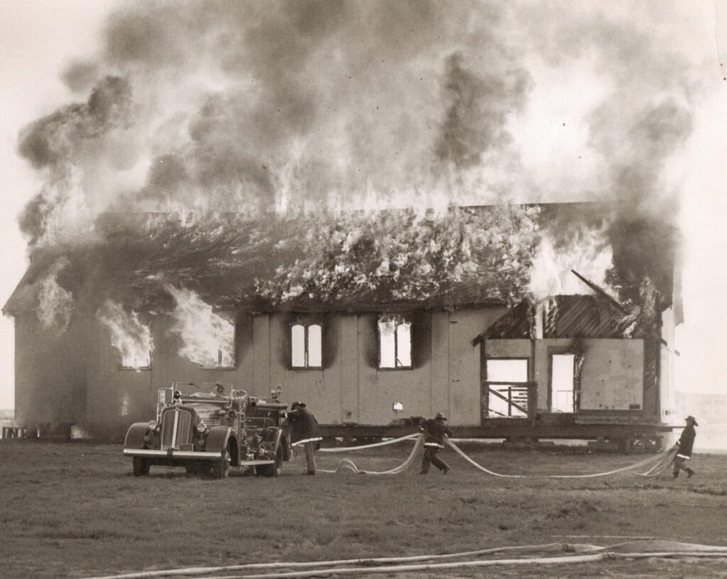 Burning building with heavy smoke; firefighters and hose; vintage fire truck in foreground.