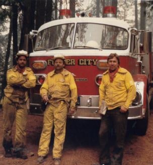 Three people in yellow uniforms stand in front of a fire truck labeled "Foster City," in a forest setting.