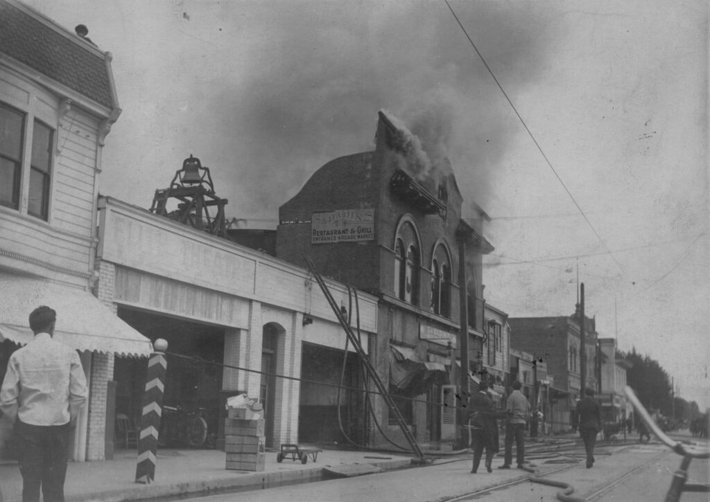 A street scene with smoke rising from a burning building as people watch; fire hoses and ladders are in use.