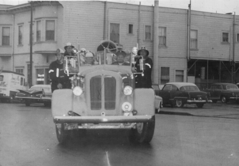 A vintage fire truck with firefighters onboard, on a street with buildings and cars in the background.