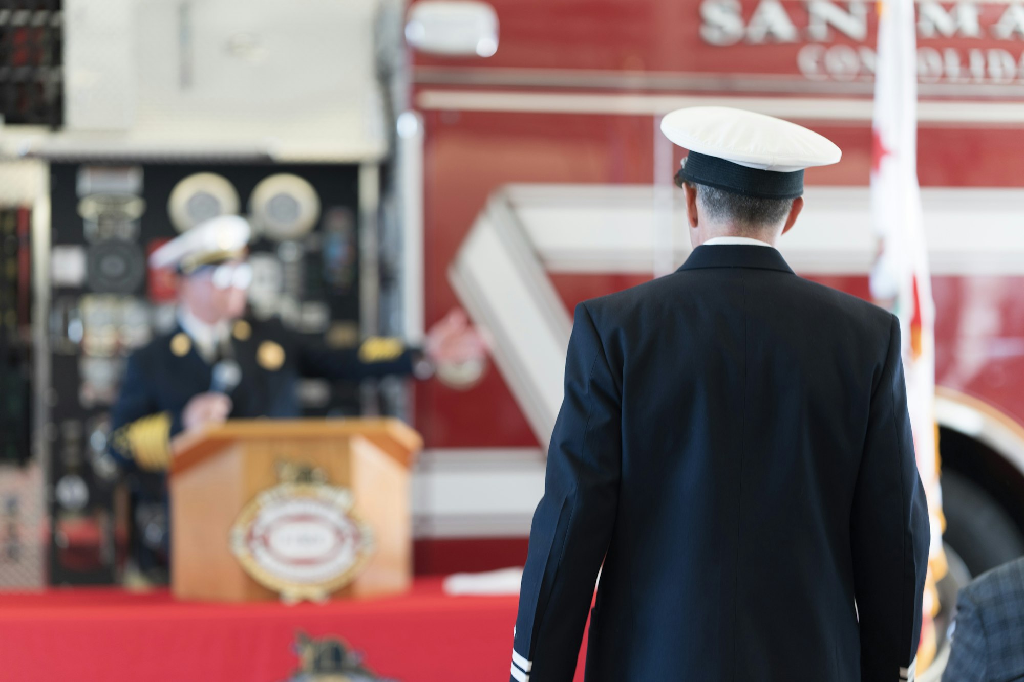 Two uniformed individuals at a podium, likely at a formal ceremony, with a red fire truck in the background.