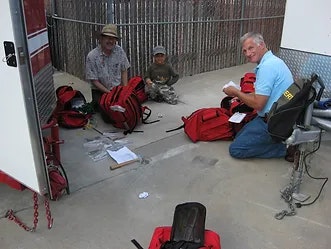 People sitting on the ground, assembling or organizing red backpacks near a trailer.