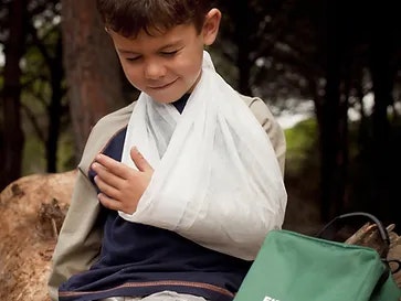A boy with a sling on his arm sits outdoors near a green first aid kit.