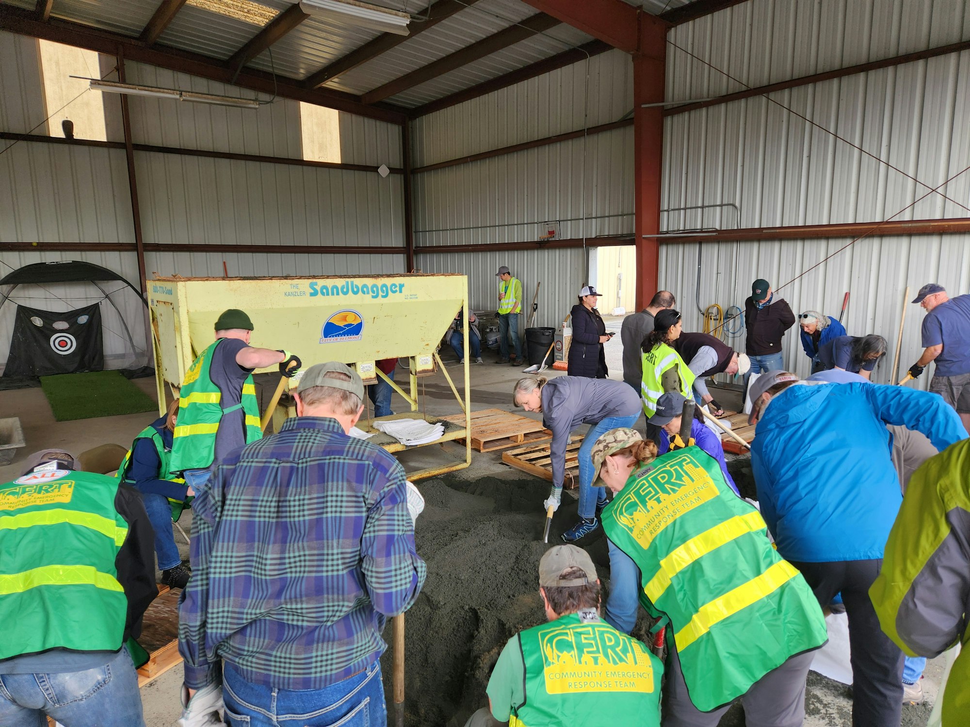 People wearing safety vests fill sandbags in a workshop setting using a sandbagging machine.