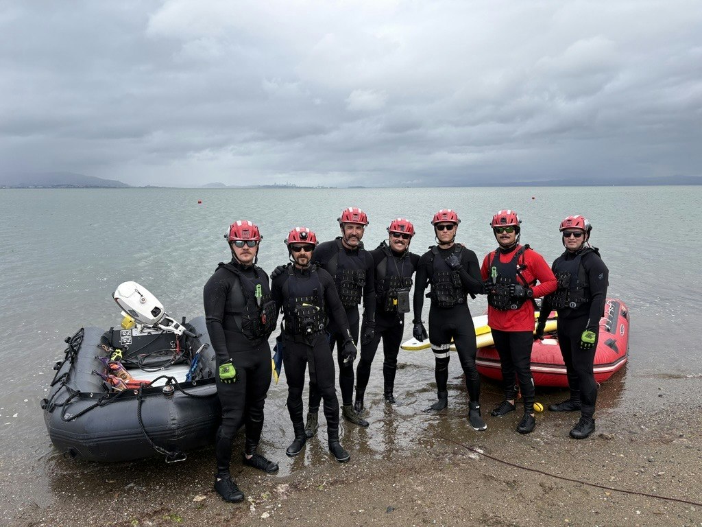 A group of six people in wetsuits and helmets standing near water, with inflatable rescue boats nearby. Overcast skies.