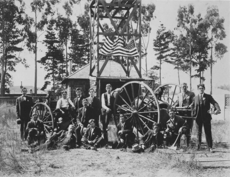 A group of men in suits pose by a wooden structure with American flags and large wheels, outdoors with trees in the background.