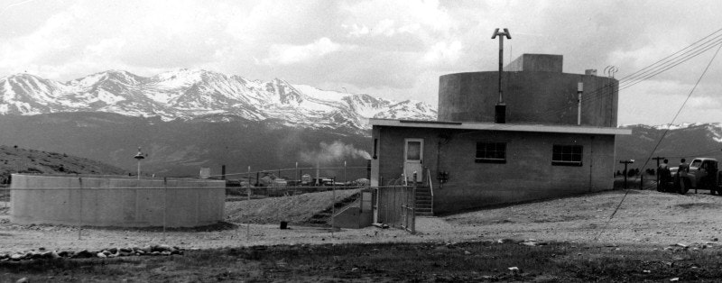 Small building with a cylindrical structure, mountains in the background, vintage black and white photo.