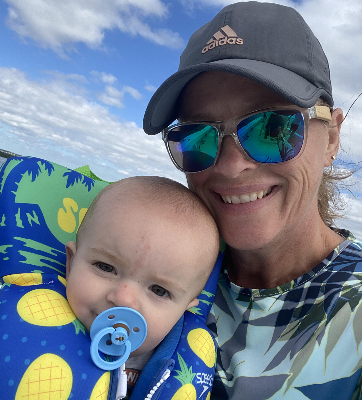 A smiling woman wearing sunglasses and a cap holds a baby in a colorful life jacket against a cloudy sky.