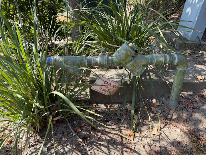 A weathered pipe system partially covered with green algae, surrounded by plants and leaves, with a warning sign beneath it.
