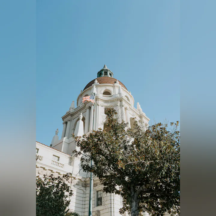 A beautiful dome of a building with a flag, surrounded by greenery, against a clear blue sky.