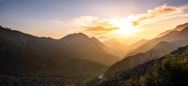 A scenic view of mountains at sunset, highlighting layers of peaks and valleys illuminated by soft, warm light.