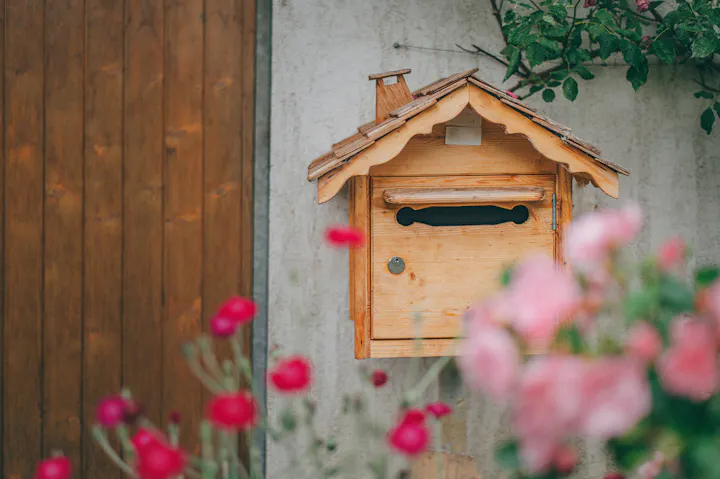 A wooden mailbox shaped like a small house, set against a wall with colorful flowers in the foreground.
