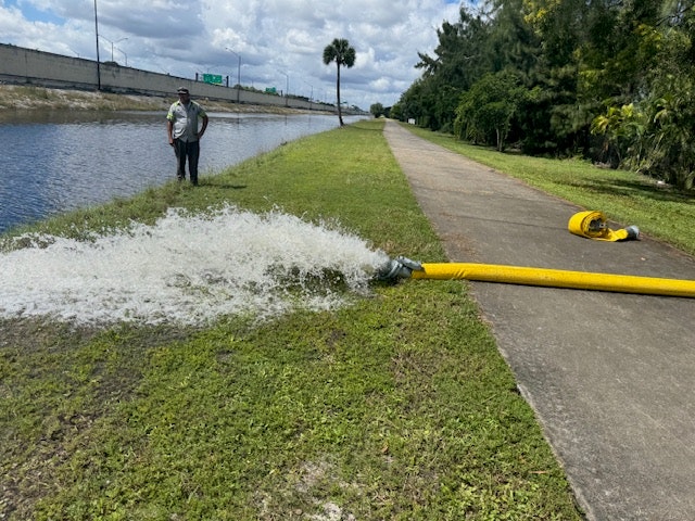 A hose is pumping water onto grass by a canal, with a person standing nearby.