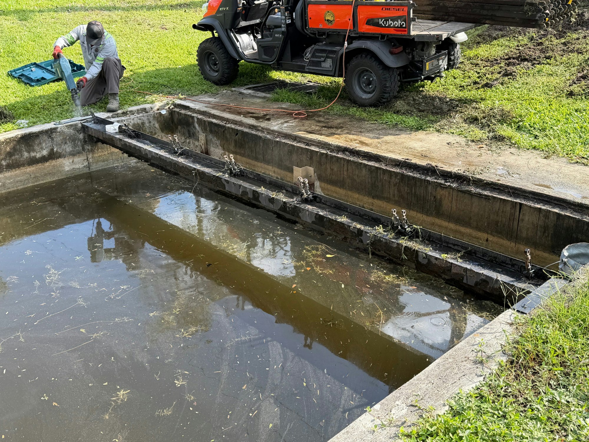 A person works near a muddy water channel, using equipment, with a utility vehicle parked nearby.