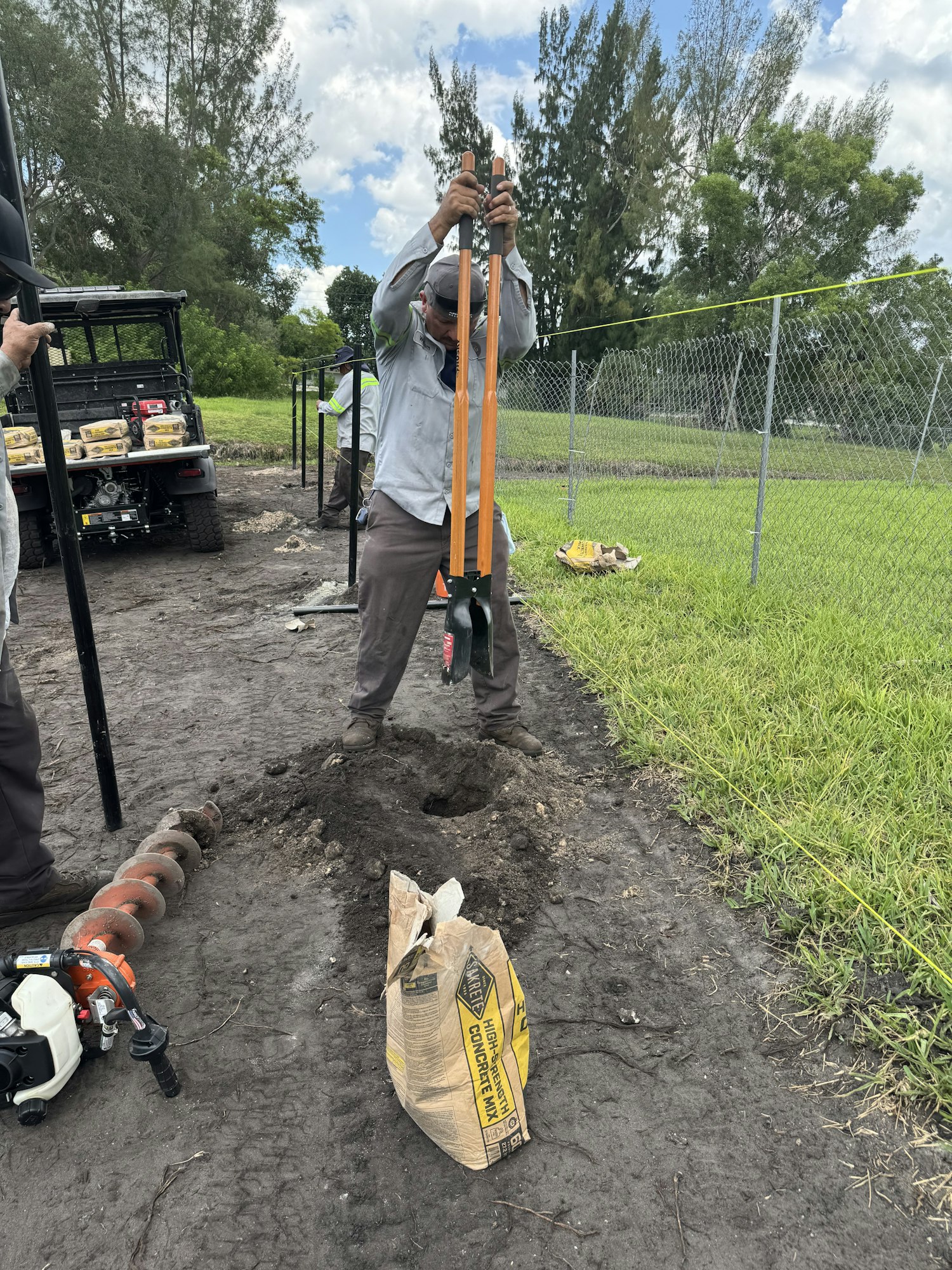 Two people are working with post hole diggers, near a bag of concrete mix, and an auger, by a fence under construction.