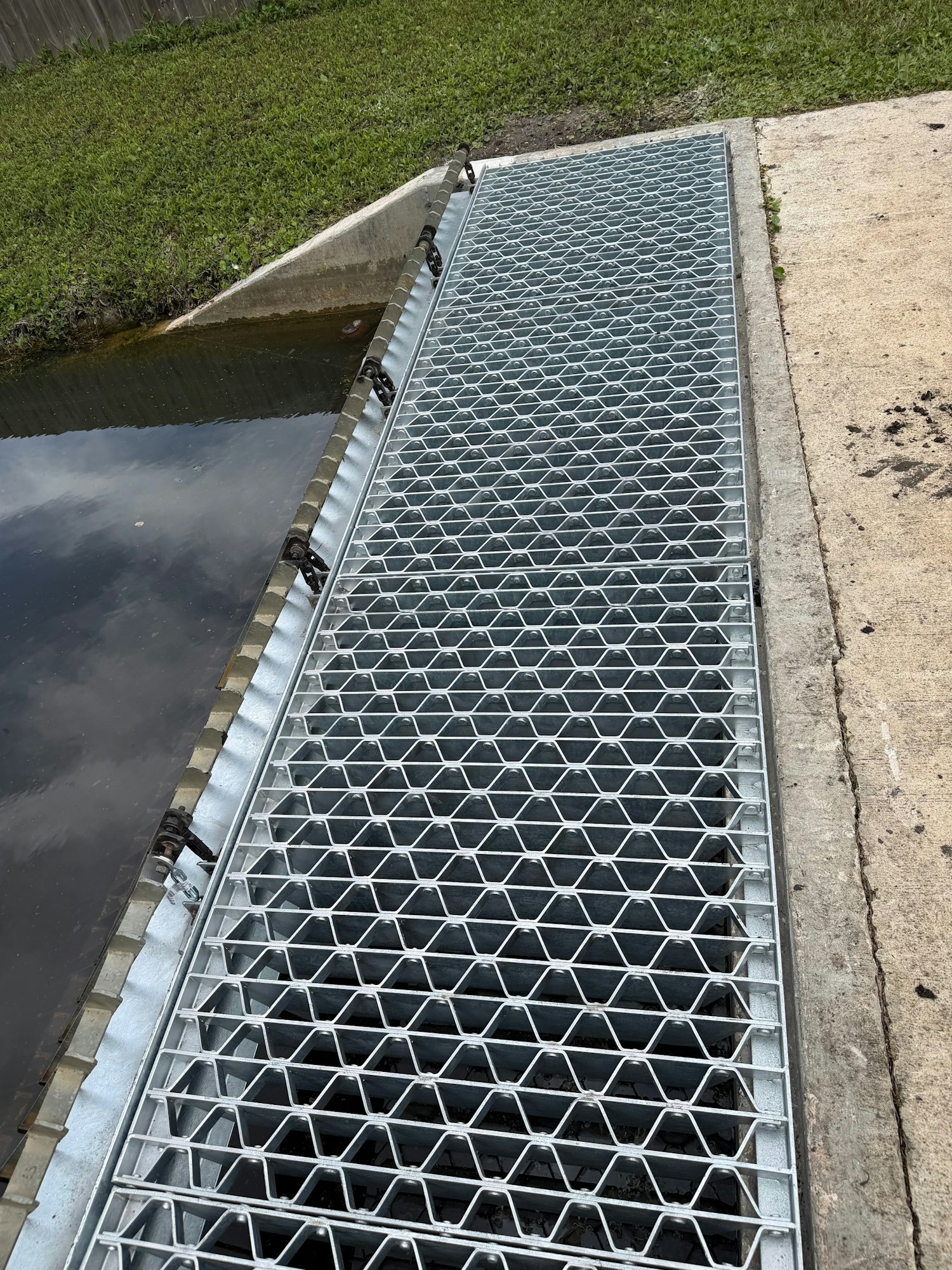 A metallic, honeycomb-patterned grate covers a waterway, set against grassy and concrete surfaces.