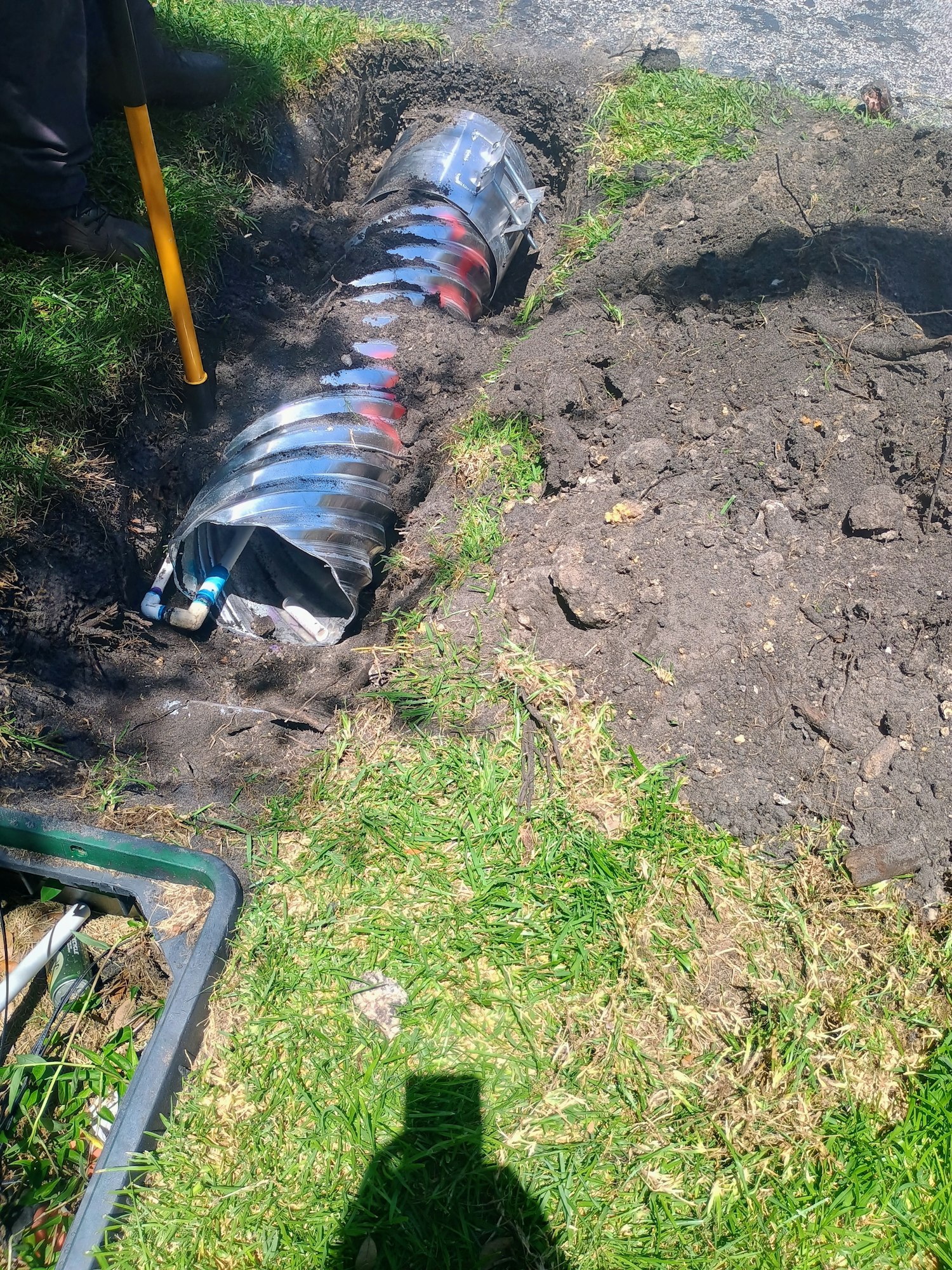 A shiny metal culvert pipe partially buried in a dug trench with soil and grass around.