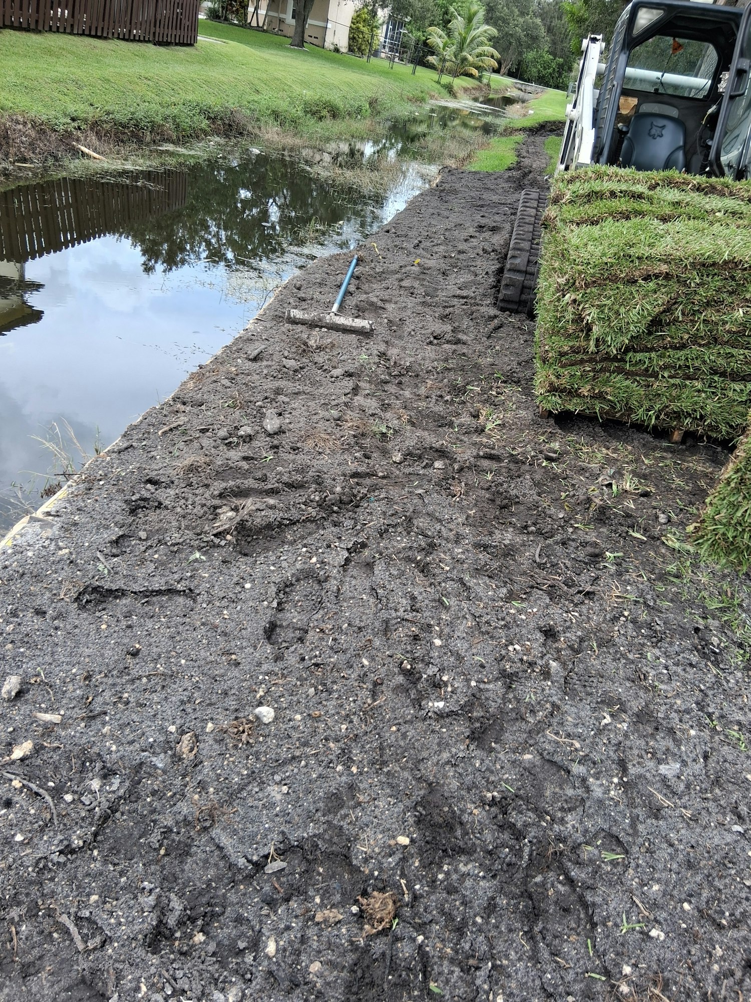 Bundles of sod on a truck near a dirt area and puddle, with a rake visible.
