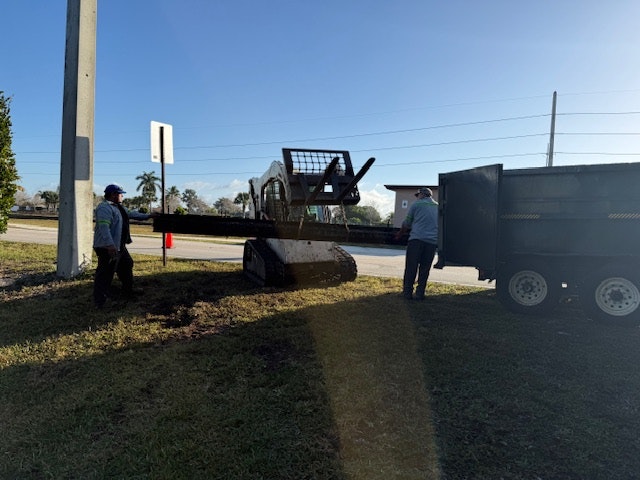 Two workers beside a skid-steer loader, unloading materials next to a parked truck, on a grassy roadside under clear skies.