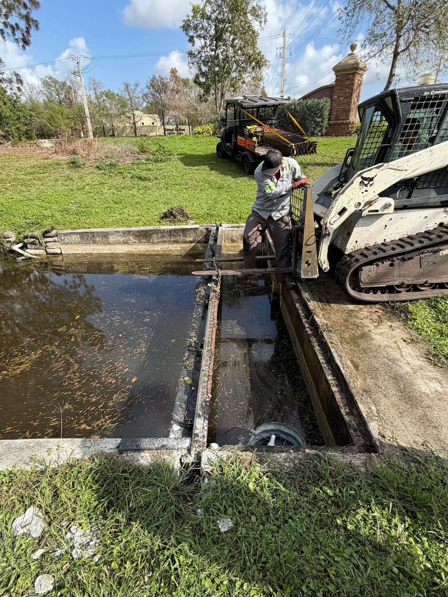 A worker is using equipment near a drainage area, while a tracked vehicle is parked nearby on grassy grounds.