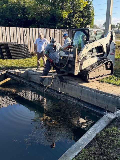A compact excavator is positioned near a canal, with two people observing and one person handling equipment near the water.