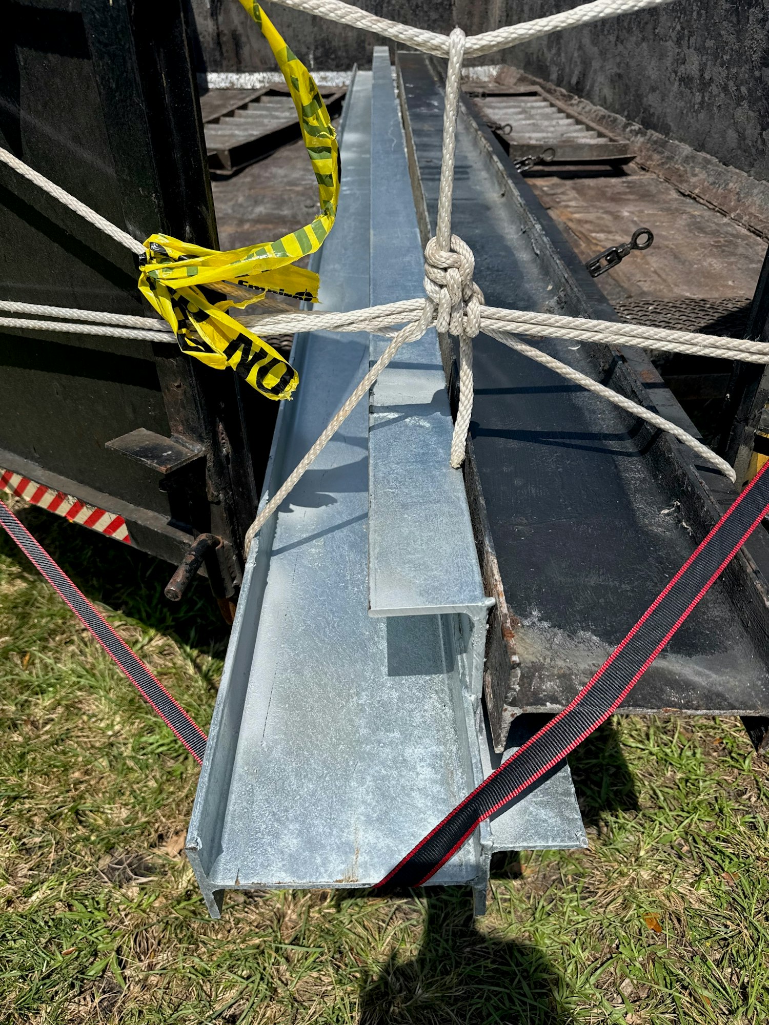 The image shows galvanized metal beams secured with ropes and straps on a surface, possibly a trailer or truck.