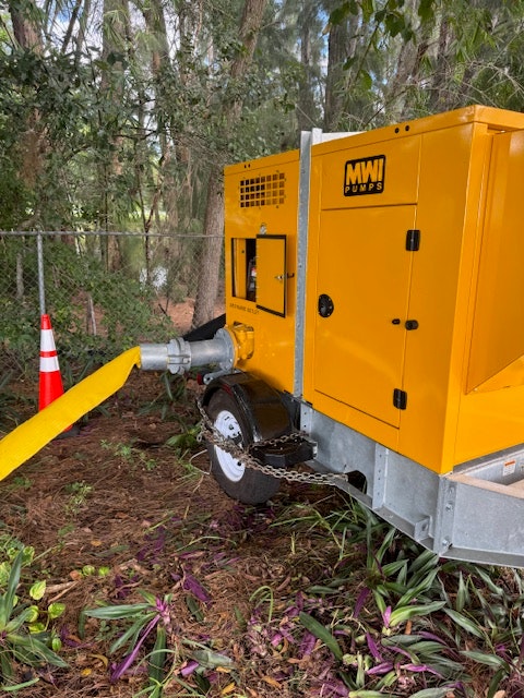 Yellow portable pump with attached hose in a wooded area, near orange traffic cone.