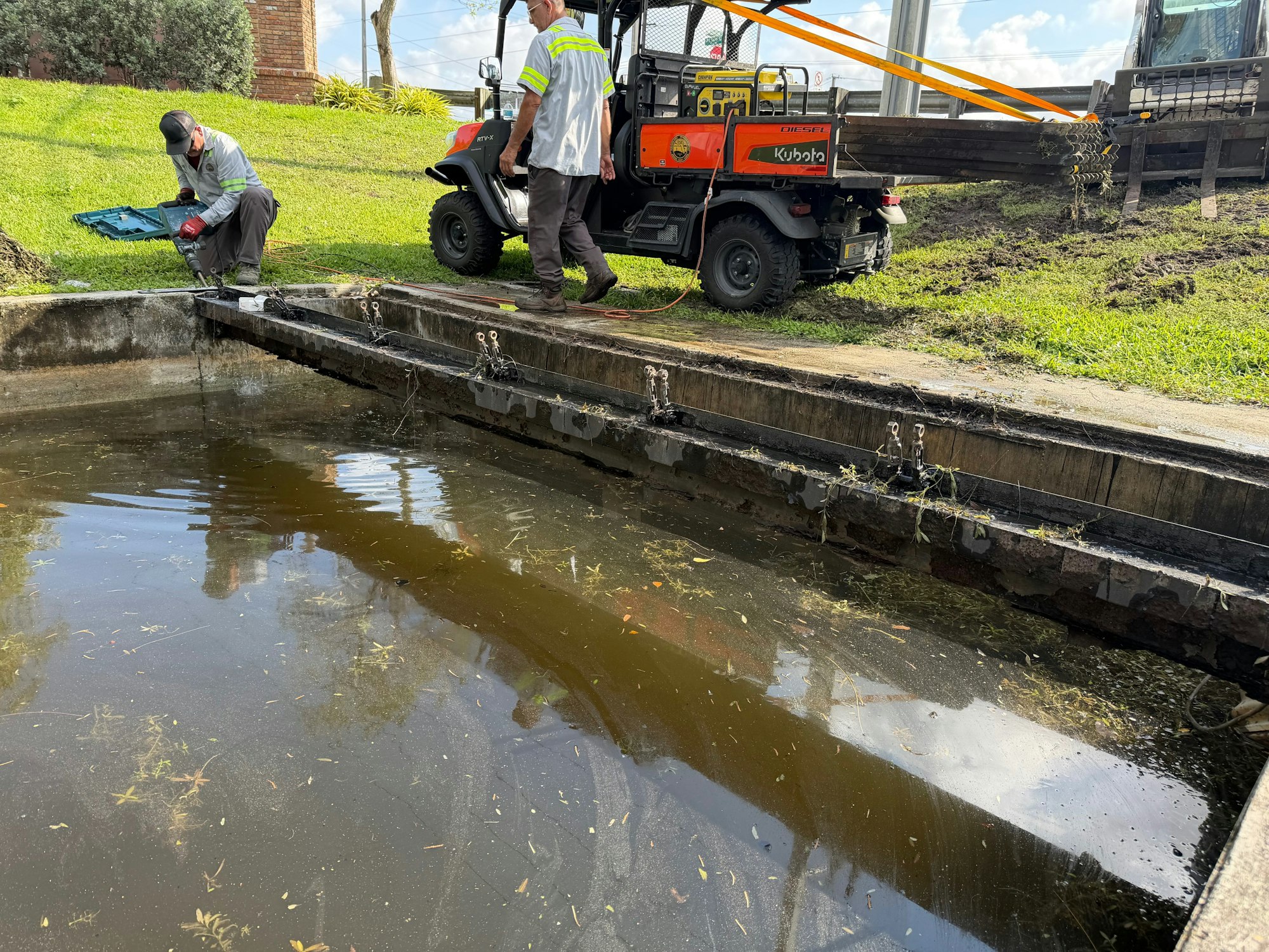 Two workers are near a muddy water tank, using equipment for maintenance while a utility vehicle is parked nearby.