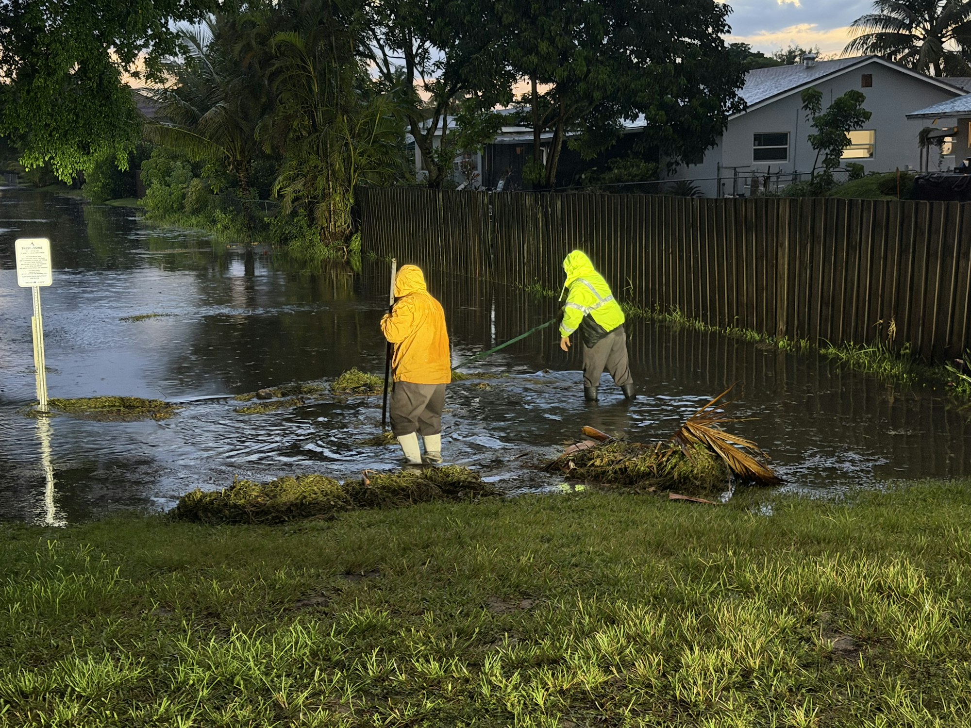 Two people in rain gear are wading through a flooded area, clearing debris from a neighborhood after heavy rain.