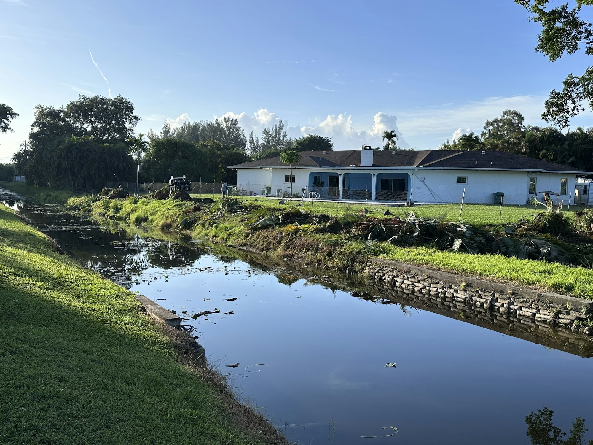 A house by a small canal with clear sky, trees, and scattered foliage on the grass.