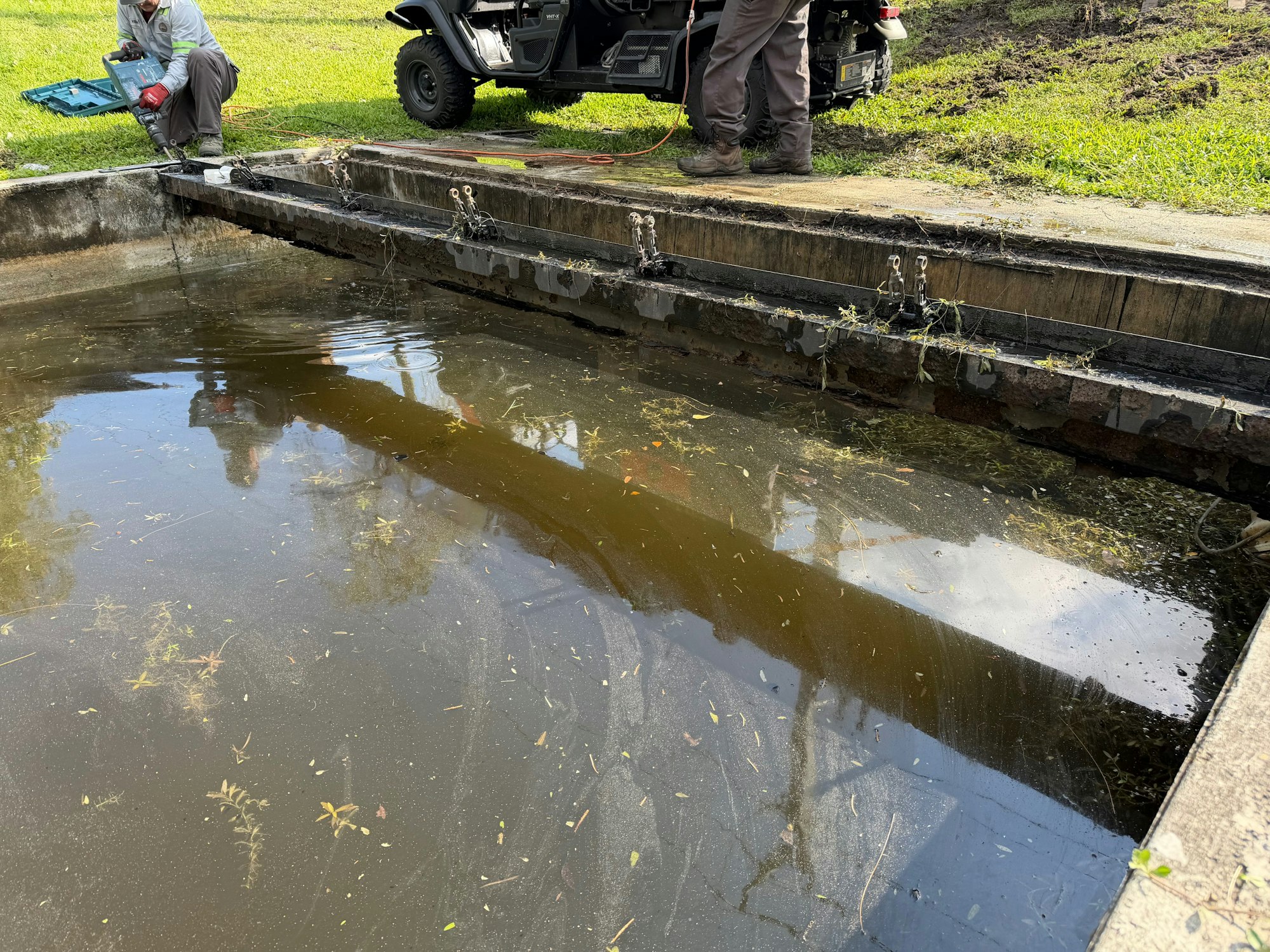 A worker is using a tool near a muddy water area with some foliage, while an ATV is parked nearby.