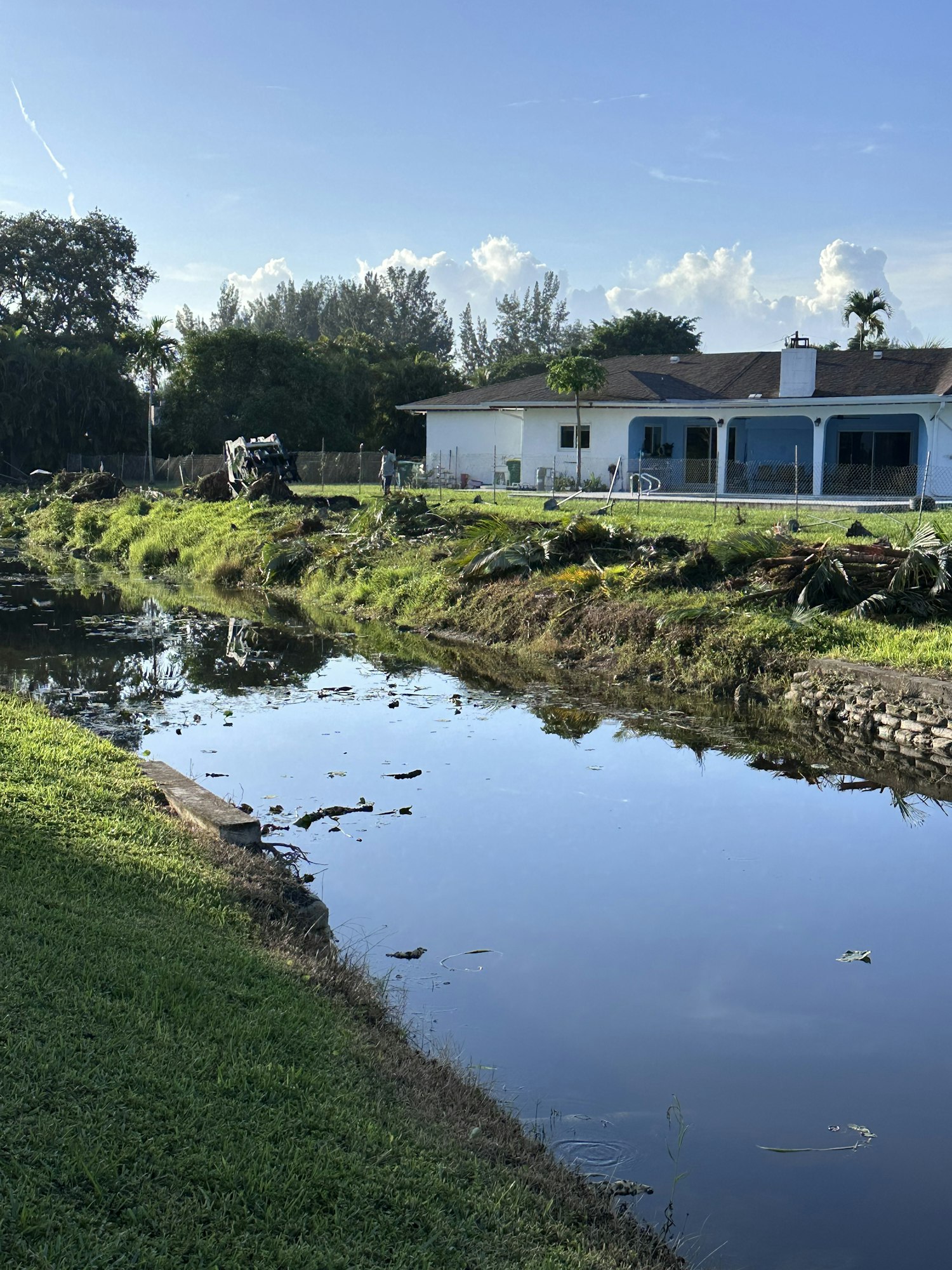 A house by a canal with a lush, grassy foreground and trees in the background under a clear blue sky.