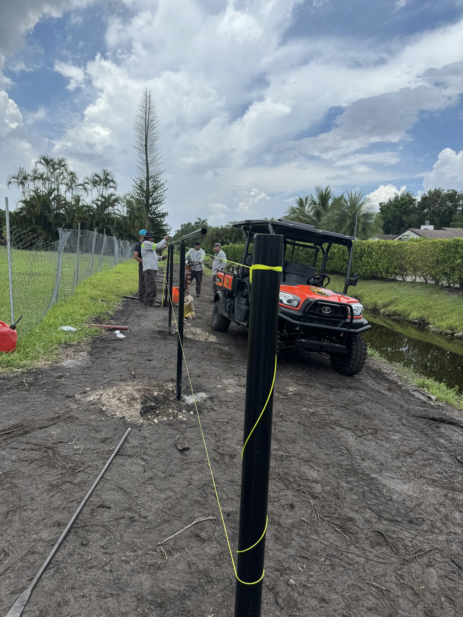 Workers install fencing near a utility vehicle, with tools and equipment on the ground, under a partly cloudy sky.