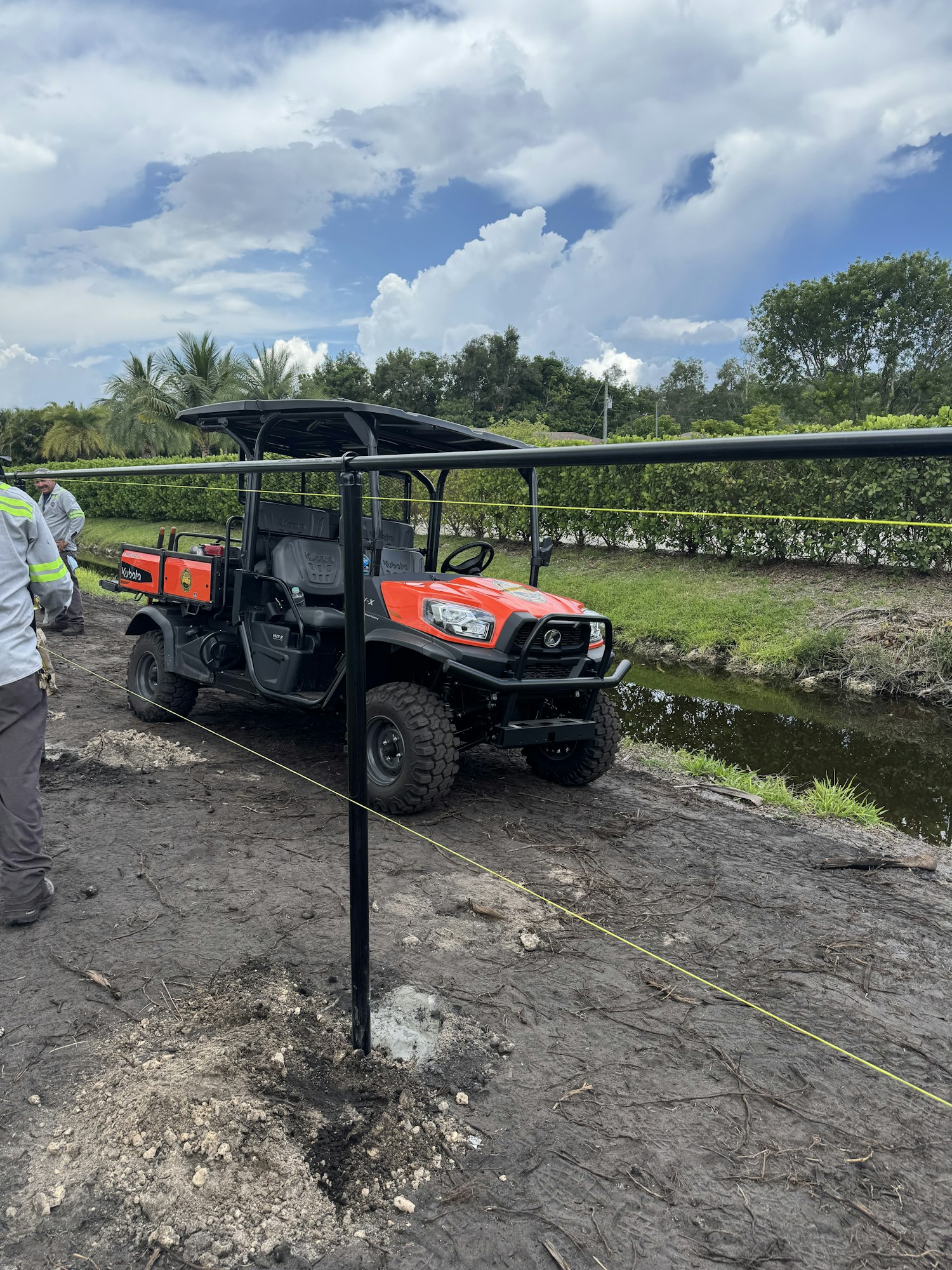 An off-road vehicle parked on dirt next to a water canal, with trees and cloudy sky in the background.