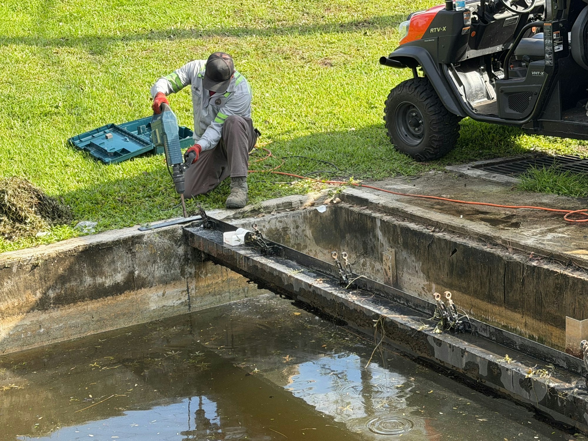 A person is using a power tool to work on a construction project near a water-filled area, with equipment and a utility vehicle nearby.
