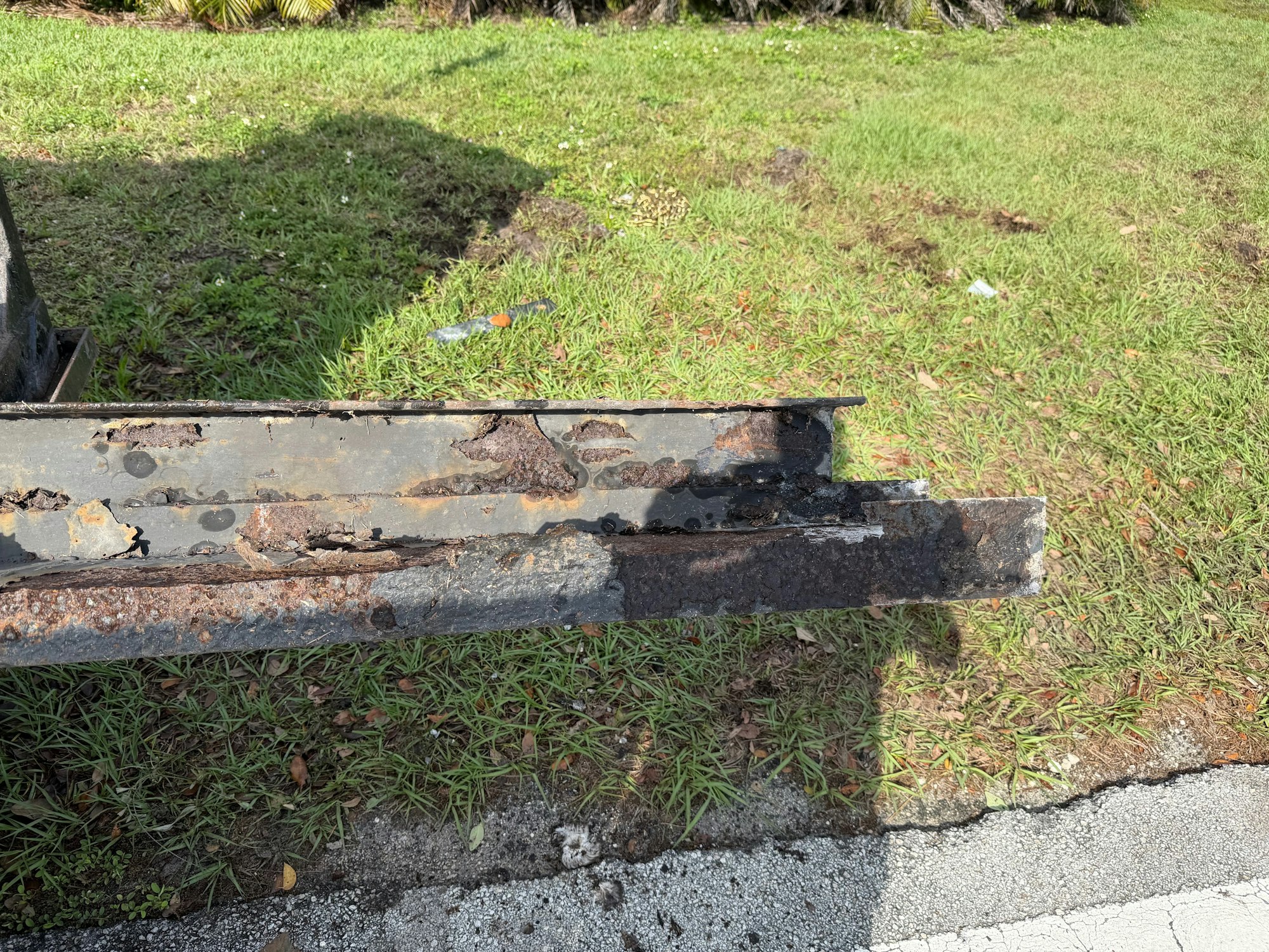 The image shows a rusted metal beam partially covered in corrosion, with grass and pavement visible in the background.